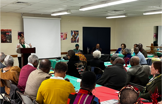 Kacey Carnegie stands at a podium speaking to a room of people sitting around large tables