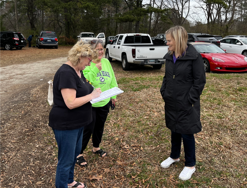 Kacey Carnegie stands in a grassy parking lot with two women, one of whom is signing a paper